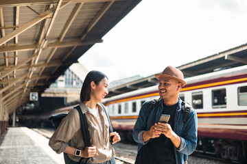 Couple Young asian traveler tourist by train Choose a trip and booking train queue by smartphone. Two Backpacker at platform train station. Happy traveller at railway on vacation holiday weekend.
