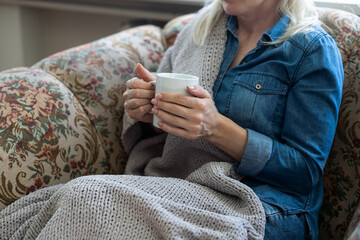 Close up of hands holding blue cup of tea or coffee. Model and cozy plaid is sitting, relax at home on sofa