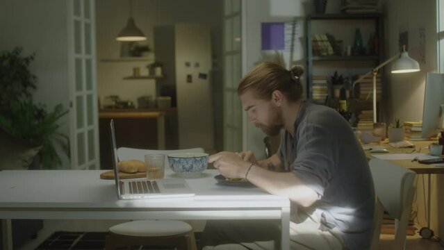 Side view of young bearded man eating breakfast and watching movie on laptop in morning at home