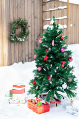 Snowy artificial fir tree decorated with Christmas ornaments and baubles outdoors in yard in wooden fence background on sunny day. Christmas and New Year concept. Copy space