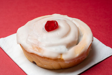 Close-up of a freshly made iced  bun with a glazed cherry on top served on a napkin. Red background.