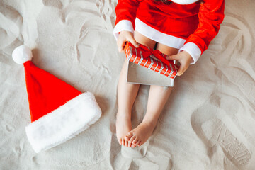 a little girl is sitting on the sand in a Santa hat. the child smiles and looks at the gift box....