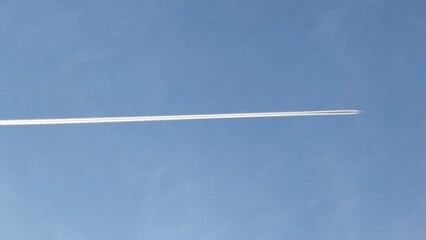 A long white trail from a flying plane. A jet plane flies in a light blue sky with barely noticeable translucent blurry clouds, leaving behind a white thermal trail.