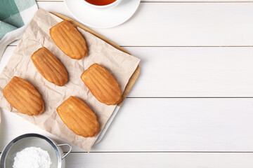 Tasty madeleine cookies, tea and powdered sugar on white wooden table, flat lay. Space for text