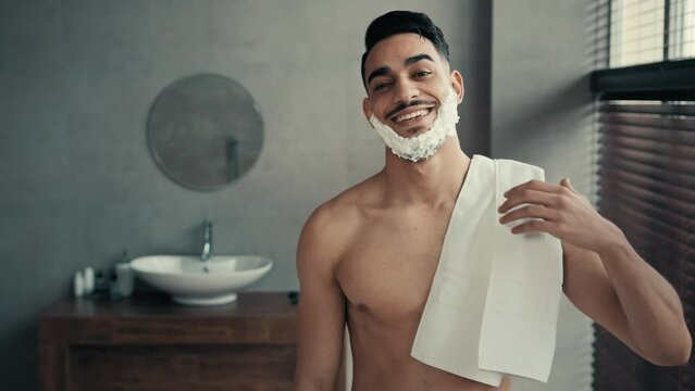 Funny male portrait in bathroom Latina guy smile posing before shave beard. Hispanic Indian smiling happy sexy man bearded guy with white soapy foam on beard shaving gel holding bath towel hygiene
