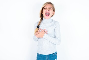 beautiful caucasian teen girl wearing gray turtleneck sweater over white wall expresses happiness, laughs pleasantly, keeps hands on heart