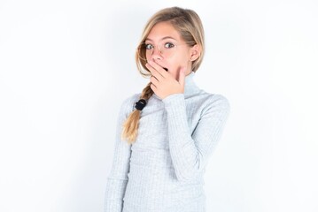 beautiful caucasian teen girl wearing gray turtleneck sweater over white wall covers mouth and looks with wonder at camera, cannot believe unexpected rumors.
