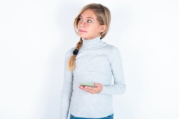 beautiful caucasian teen girl wearing gray turtleneck sweater over white wall holds mobile phone uses high speed internet and social networks has online communication. Modern technologies concept