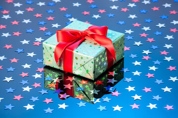 Single green gift box with red ribbon bow on blue reflective surface with glitter stars. Gift box...
