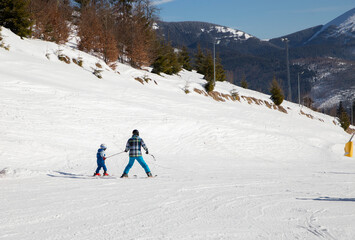 back of a child and an adult in warm overalls, on skis. skiing lesson on a snowy slope in the mountains, playing sports with dad, an instructor. Ski training. Sports education. Active winter holidays