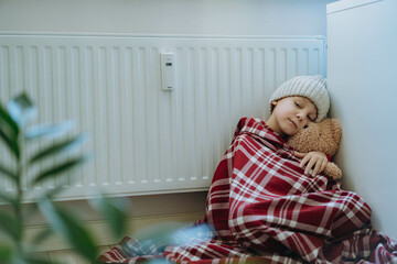 cute little boy wrapped id plaid wearing knit hat sitting by heater hugging teddy bear, cold at home