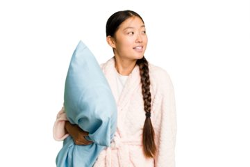Young asian woman wearing a pajama holding a pillow isolated looks aside smiling, cheerful and pleasant.