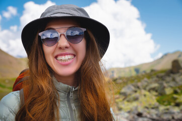 Smiling young girl against a blue cloudy sky and mountains. Portrait of a happy woman tourist in sunglasses and panama