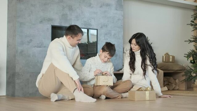 Family with young son sit on floor near fireplace in morning and give open gifts. Happy children receive gift boxes for holiday. Happy family spending time together at Christmas time