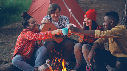 Joyful young people friends are clinking glasses with drinks sitting around fire nera tent in...