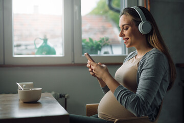 side view of caucasian pregnant woman with headphones sits in kitchen and using mobile phone