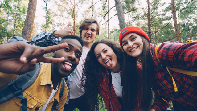 Multiracial group of friends travelers is taking selfie in wood looking at camera, posing with funny faces and trendy gestures. Millennials are wearing modern tourist outfit.