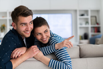 Couple Watches TV together while Sitting on a Couch in the Living Room. Girlfriend and Boyfriend embrace, cuddle, talk, smile and watch Television Streaming Services. Home with Cozy Stylish Interior.