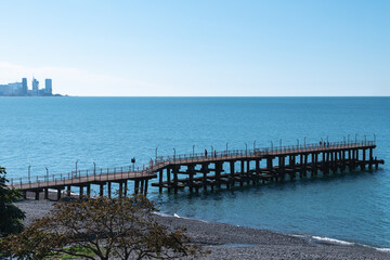 Fototapeta premium A wooden jetty located diagonally near the rocky shore against a clear blue sky and the modern city of Batumi in the distance