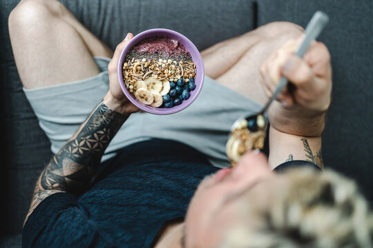 Young adult man with tattooed arm eating muesli bowl after workout while sitting on the couch. Healthy lifestyle and wellbeing