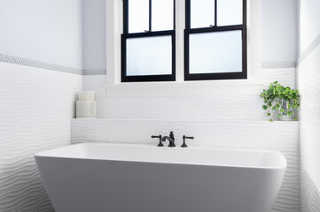 Fototapeta na wymiar A gorgeous bathroom with a standalone bathtub and black faucet surrounded by custom white tile. 