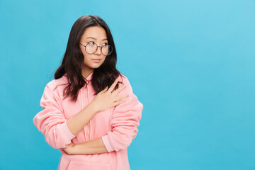 Fototapeta premium Confused sad cute Asian young lady in pink hoodie sweatshirt wear round glasses looks aside posing isolated on over blue studio background. The best offer for ad. Eyewear for vision correction concept