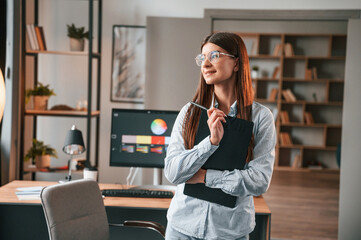 Standing against pc and holding notepad. Young female designer is in the modern office