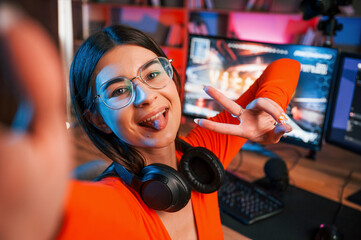 Smiling, making victory gesture and doing selfie. Young beautiful game streamer is indoors with pc
