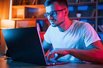 Positive emotions. Man in glasses and white shirt is sitting by the laptop in dark room with neon lighting