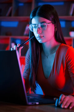 Holding stylus. Beautiful woman in glasses and red wear is sitting by the laptop in dark room with neon lighting