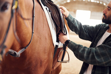 Process of preparation the animal for a ride. Young man with a horse is in the hangar