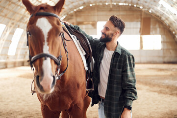 Standing with animal and smiling. Young man with a horse is in the hangar