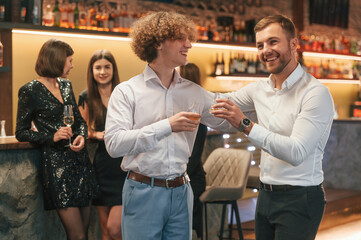 Two men are standing together in formal clothes on the party event. Friends are behind