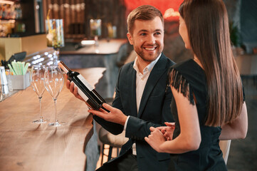 Smiling and holding bottle of wine. Man and woman are sitting at the bar with alcohol drink