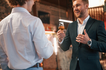 Two men are standing at the party together with alcohol drinks in hands