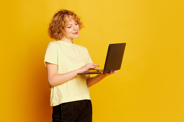 Portrait of young smiling girl, student in white t-shirt with laptop isolated on yellow color background. Concept of beauty, fashion, emotions, studying