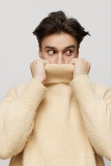 Vertical studio shot. a cute dark-haired young man stands in a beige turtleneck on a light gray background and smiles pleasantly, pointing his hands in different directions.