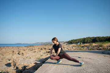 Fototapeta na wymiar Woman in sportswear doing stretching exercise at seaside. Female practicing workout outdoors during sunny day