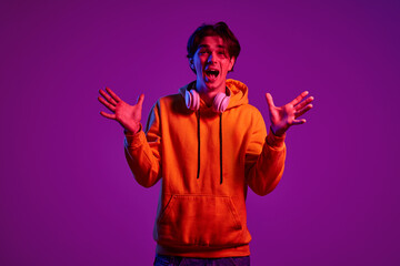 Portrait of young man in casual hoodie posing with headphones isolated over purple background in neon light. Excited and happy