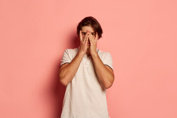 Portrait of young man in casual white T-shirt posing, covering eyes in disappointment pink...