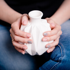 Female hands holding white ceramic vase. Manicure with pink and gray color nail polish, square photo