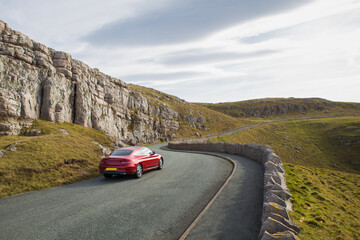Fototapeta premium Red car in the street taking a turn with background of rocky mountains.