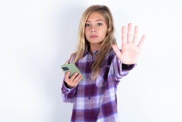 beautiful caucasian teen girl wearing plaid purple shirt over white wall using and texting with smartphone with open hand doing stop sign with serious and confident expression, defense gesture