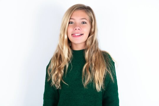 Funny beautiful caucasian teen girl wearing green knitted sweater over white wall makes grimace and crosses eyes plays fool has fun alone sticks out tongue.