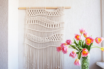 Stylish macrame wall hanging on the white wall above the chest of drawers. Cozy  interior of living room with  flowers in vase, macrame decor. Minimalistic concept of home decor. - obrazy, fototapety, plakaty