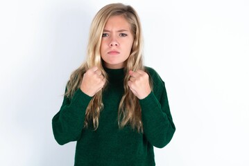 Displeased annoyed beautiful caucasian teen girl wearing green knitted sweater clenches fists, gestures pissed, ready to revenge, looks with aggression at camera stands full of hate, being pressured