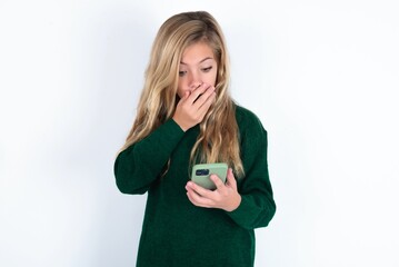 beautiful caucasian teen girl wearing green knitted sweater over white wall being deeply surprised, stares at smartphone display, reads shocking news on website, Omg, its horrible!