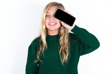 Fototapeta premium beautiful caucasian teen girl wearing green knitted sweater over white wall holding modern smartphone covering one eye while smiling