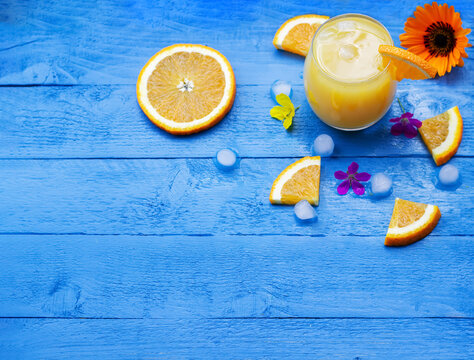 Flat Lay Of Fresh Orange Juice In A Glass With Slices Of Ripe Oranges, Ice Cubes And Flowers On The Blue Wooden Table