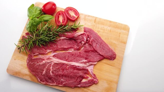 Raw beef steak on wooden cutting board with basil, rosemary and cherry tomatoes isolated on white, dolly shot